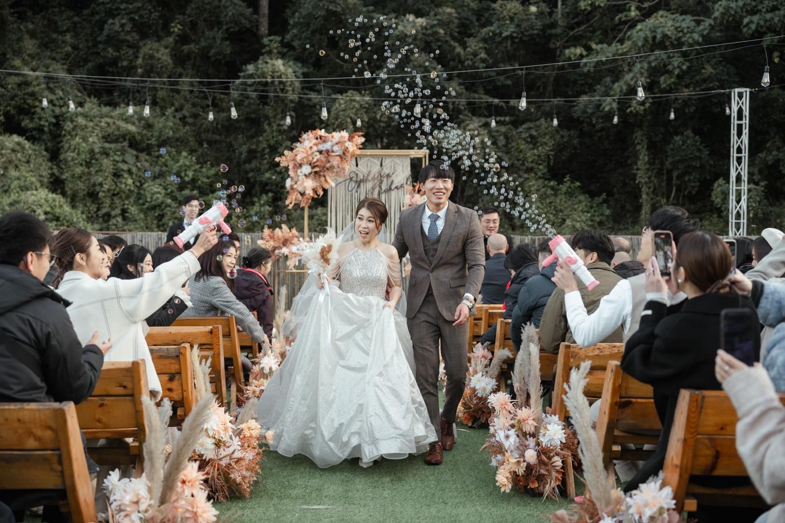Couple walking down aisle after wedding ceremony, guests cheering. Outdoor setting with floral decor.