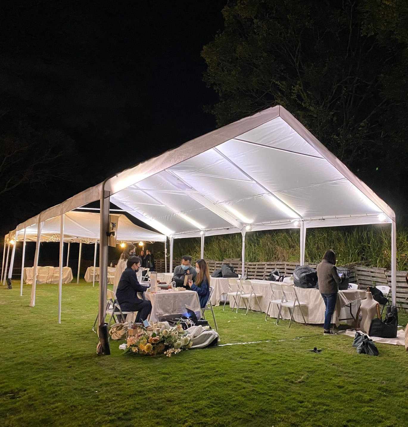 Outdoor event under a lit white tent at night. People sit at tables, grass lawn visible.