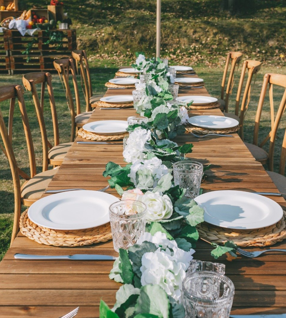 Long wooden table set for dining outdoors, decorated with white flowers and greenery.