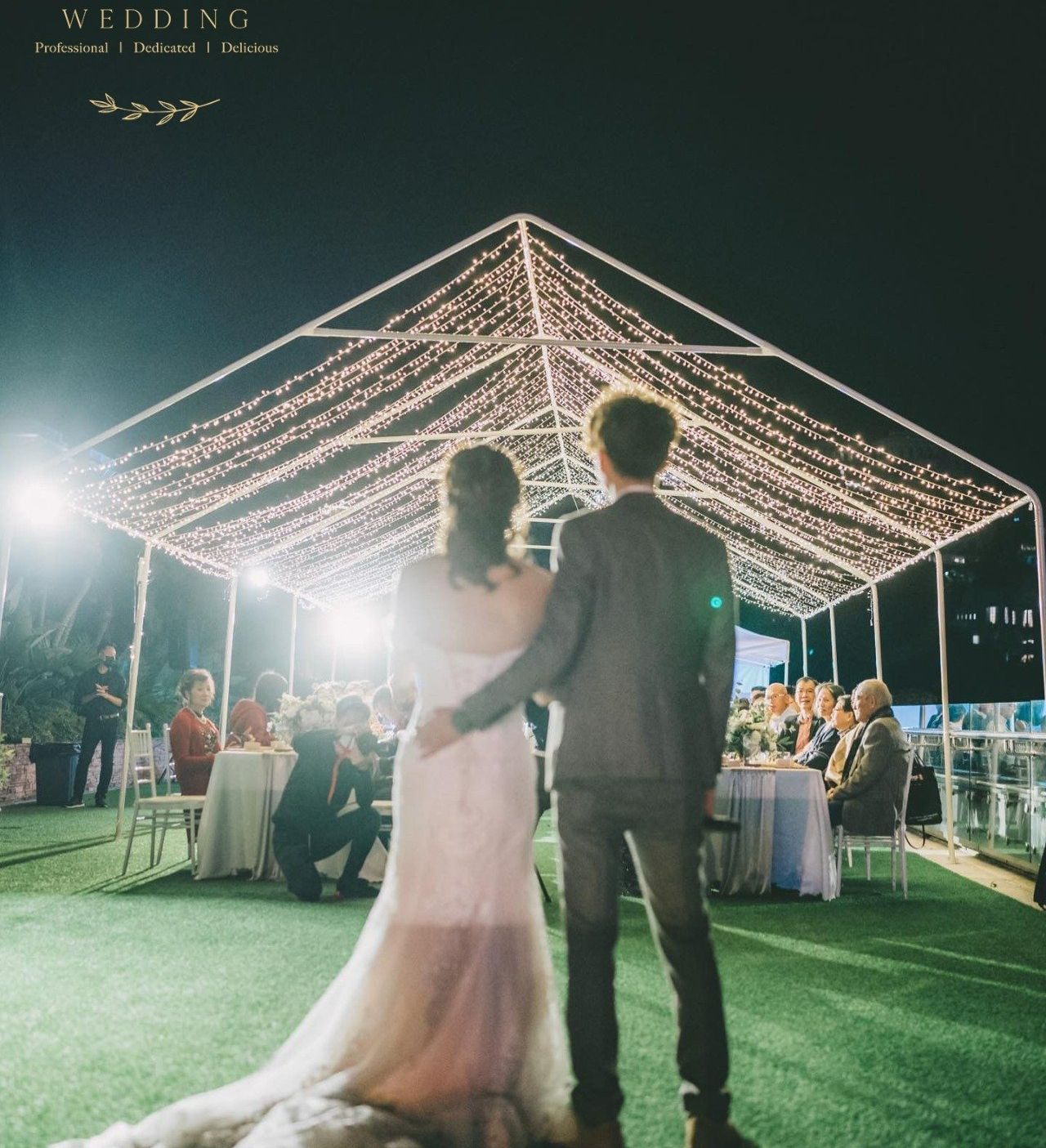 Wedding couple under a string light canopy, embracing, overlooking guests at a reception. Nighttime outdoor setting.