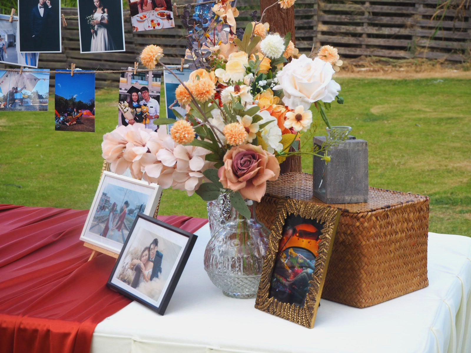 Wedding display with flowers, photos, and a framed painting on a table with a red runner and wicker box.