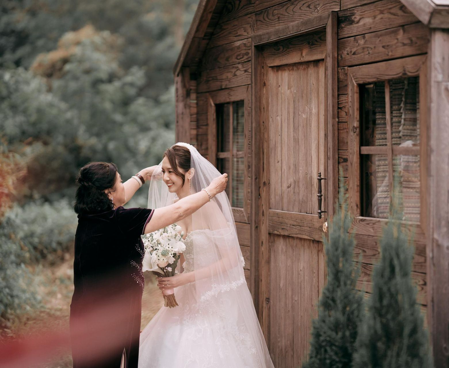 Bride adjusts her veil with help from another person near a wooden structure, holding a bouquet, outdoors.
