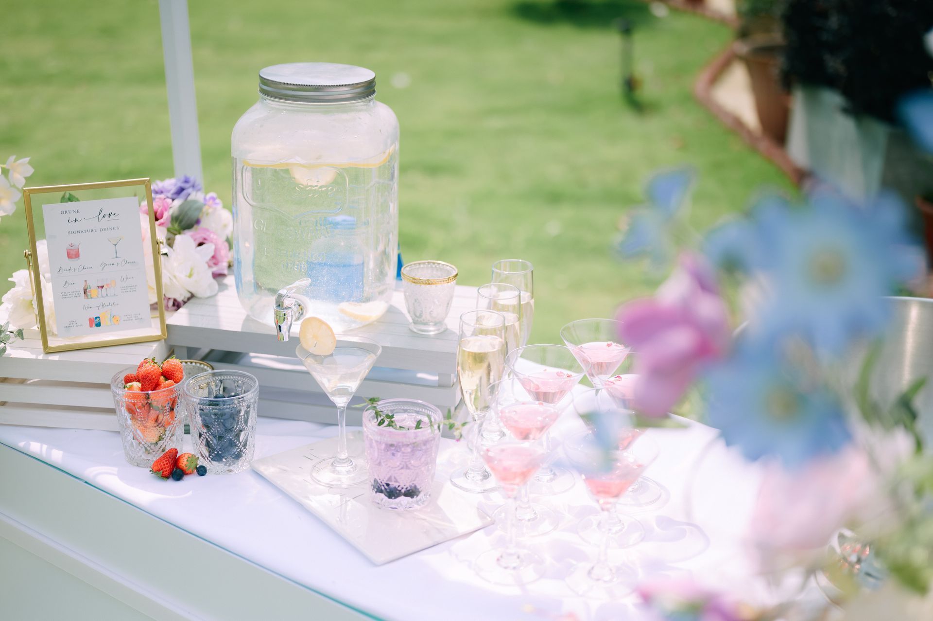 Pastel outdoor dessert table with glass jars, flowers, and candles on a white tablecloth