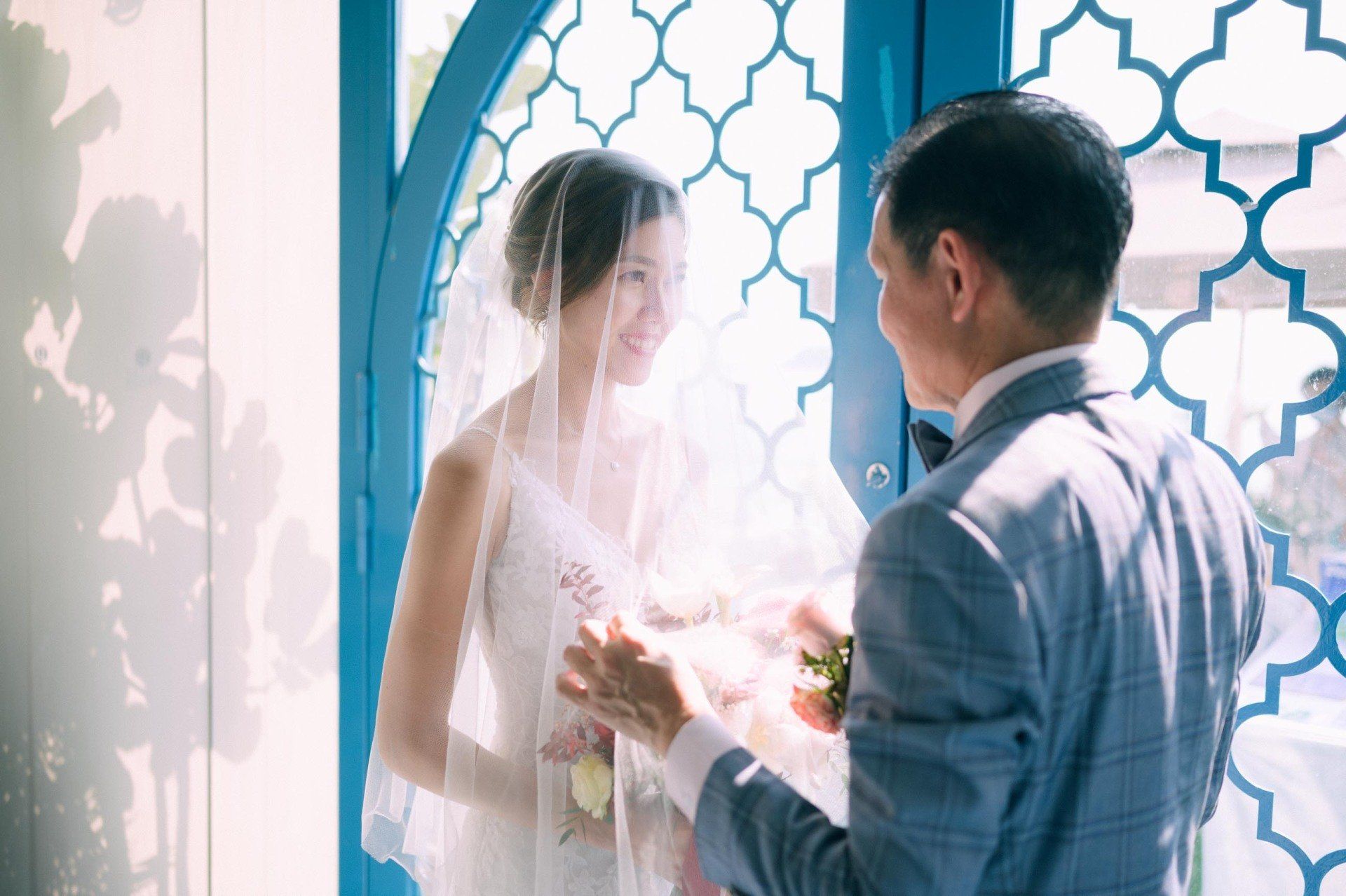 Bride smiling at groom, who is lifting her veil in front of a blue decorative window.