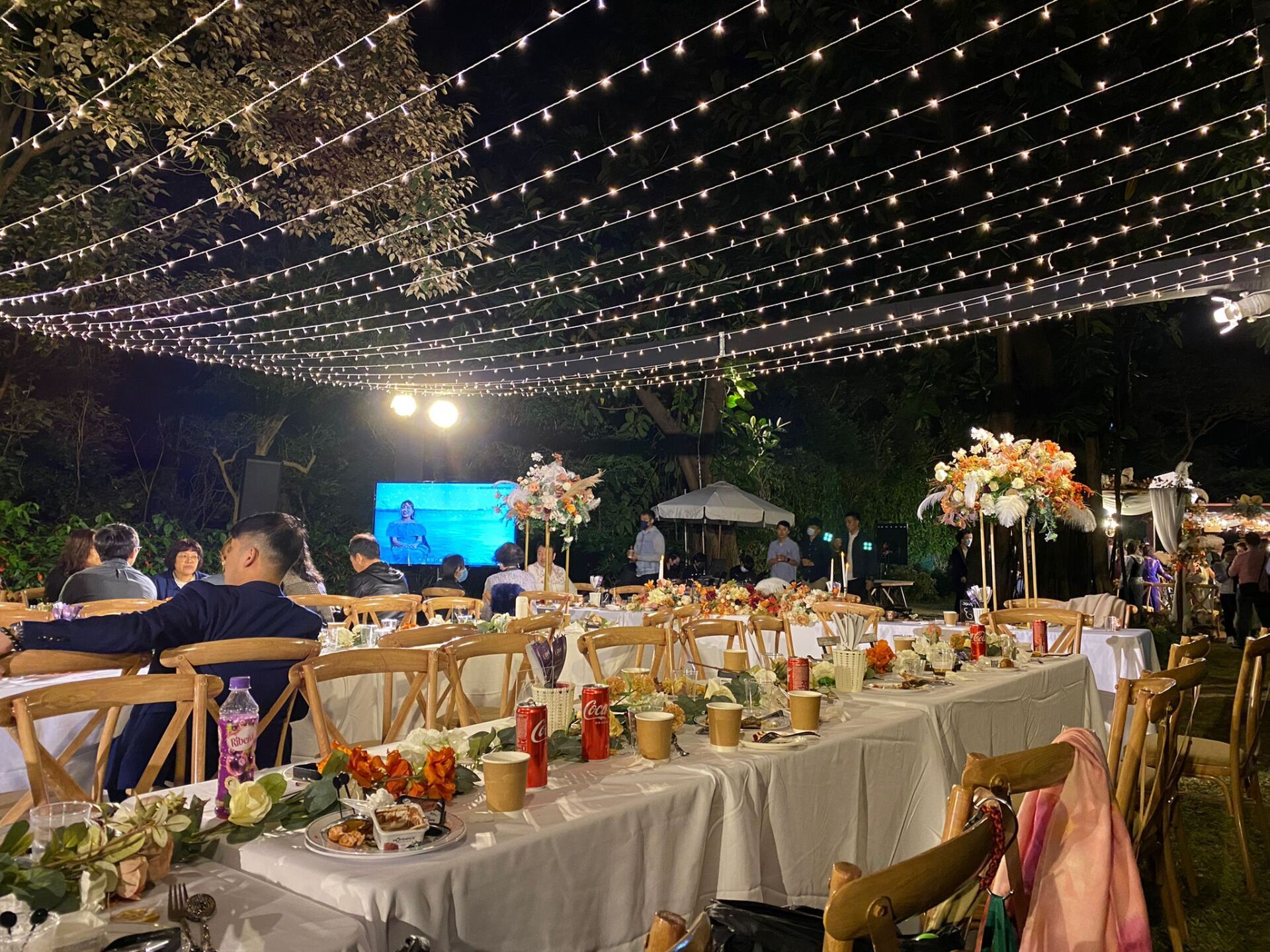 Outdoor nighttime event with long tables, string lights, and a stage.