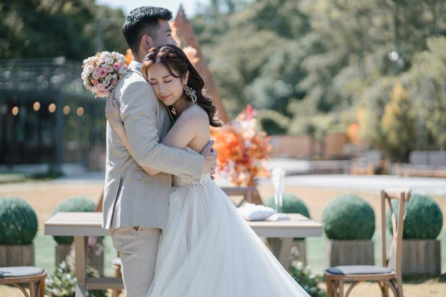Bride and groom embrace outdoors; she in a white gown, he in a tan suit; wedding setting with floral decor.