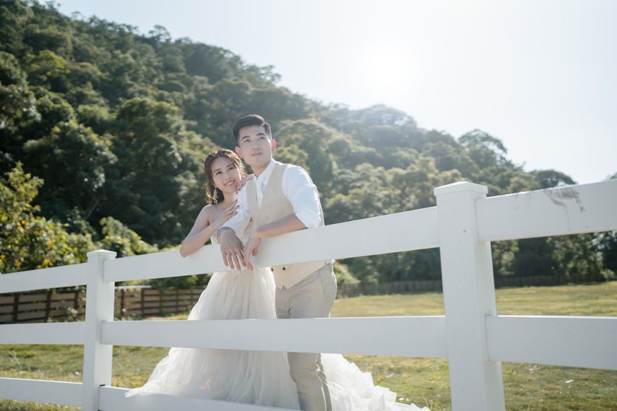 Couple in wedding attire leaning on a white fence, green hills in background.