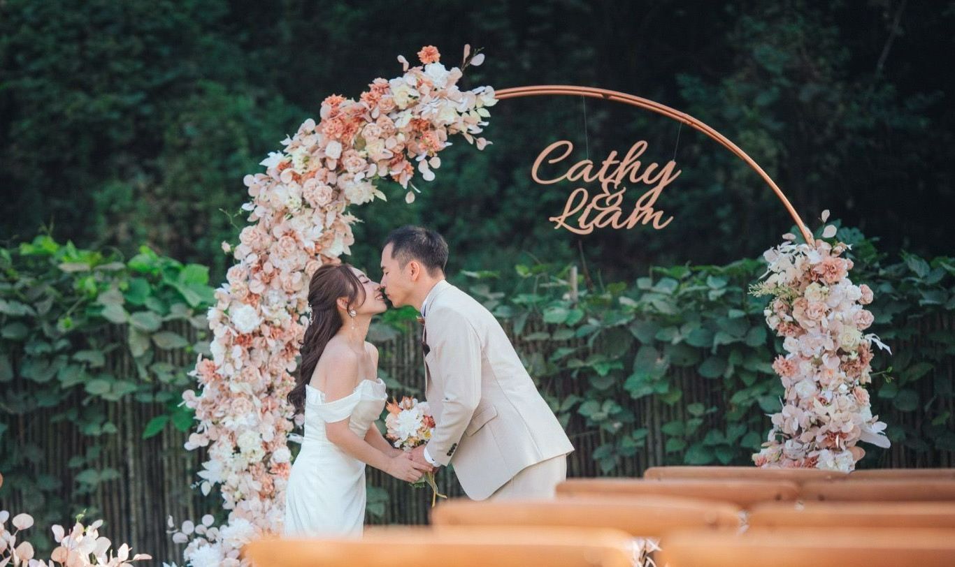 Couple kissing under floral archway at outdoor wedding.