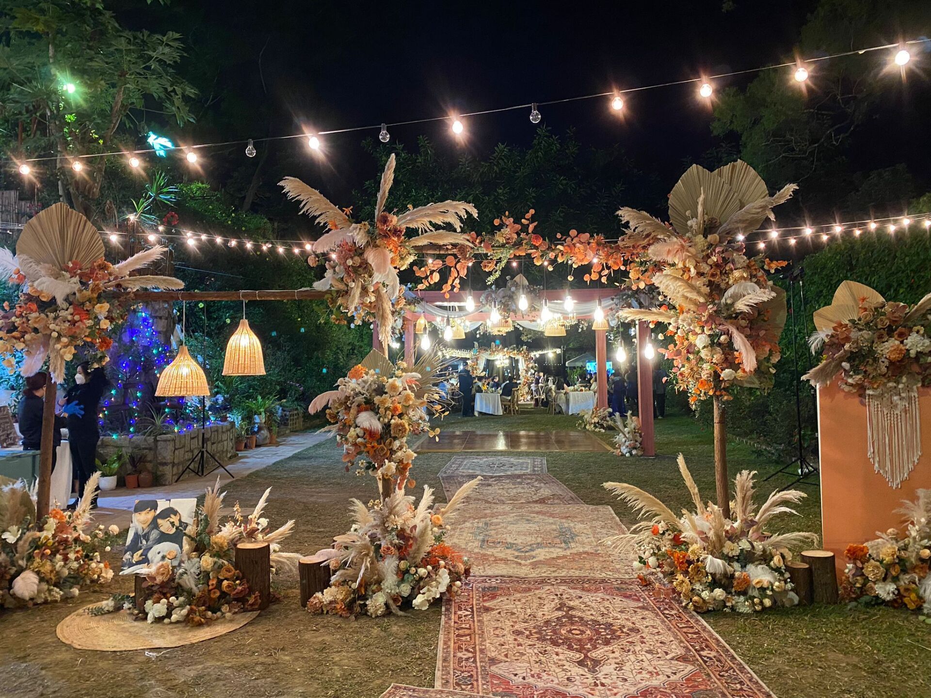 Outdoor wedding reception with floral archway, string lights, and rug.