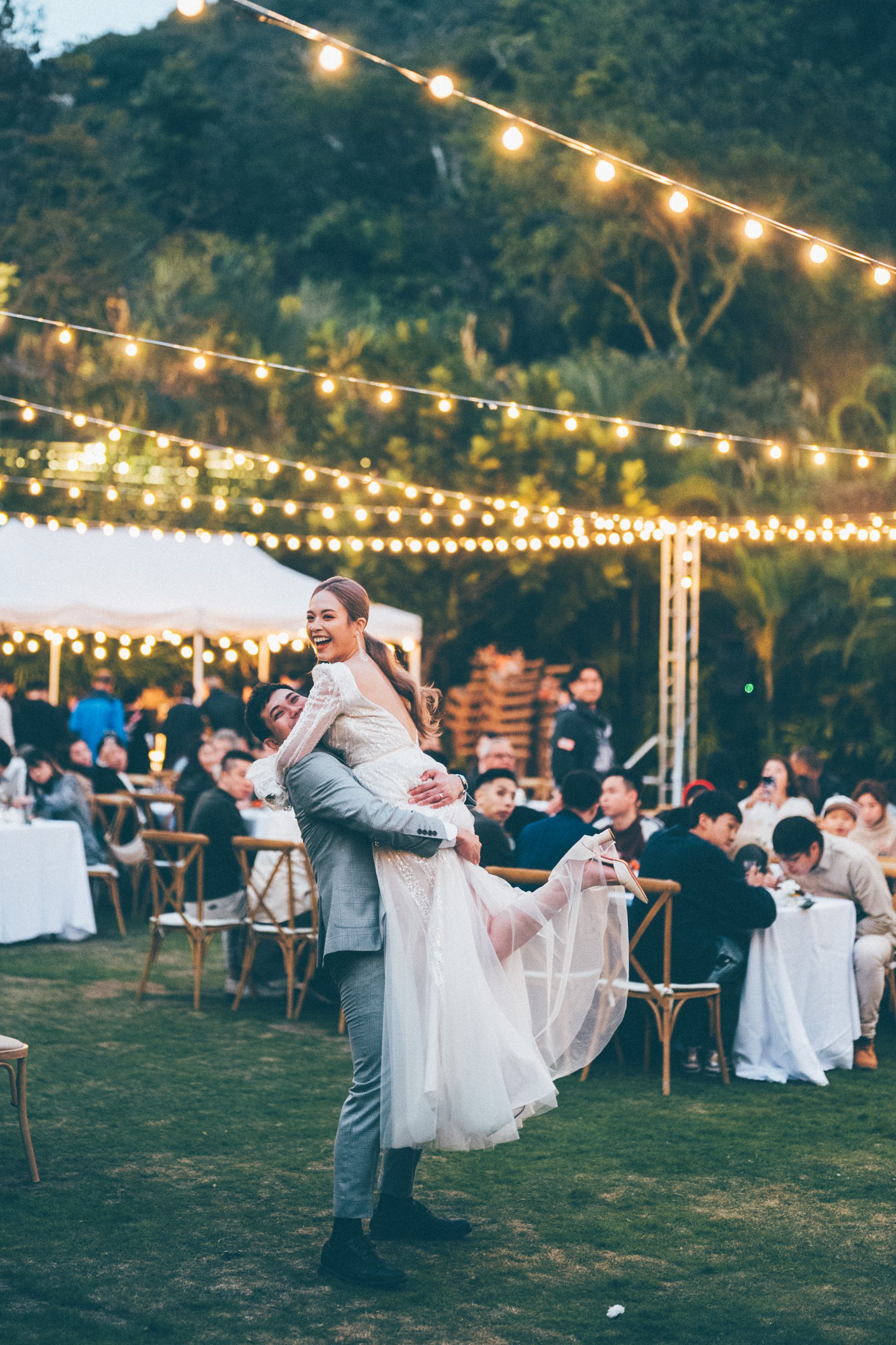 Groom carries bride at outdoor wedding reception, smiling, under string lights.