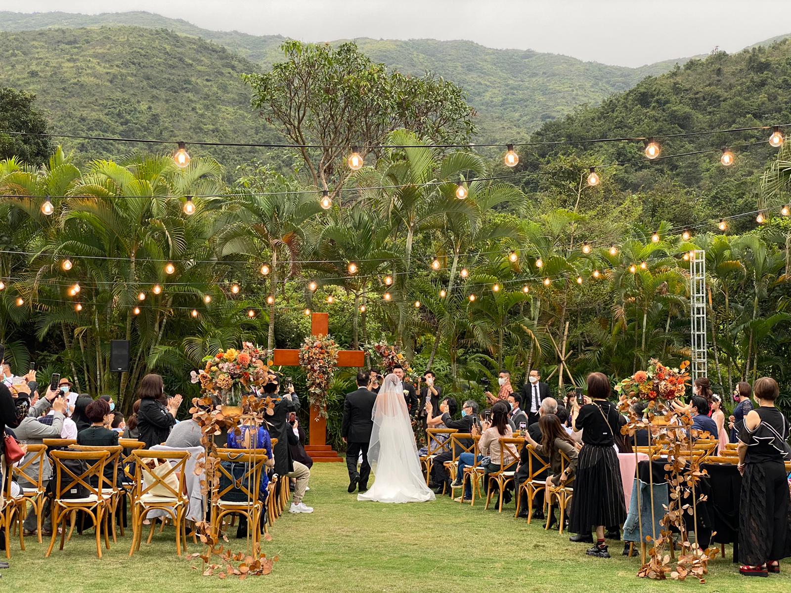Wedding ceremony outdoors with couple walking toward a cross-shaped altar, guests seated, string lights, greenery.