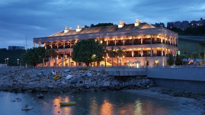 Lit-up multi-story building by water at dusk. Brown roof, windows with interior lights. Rocky shoreline.