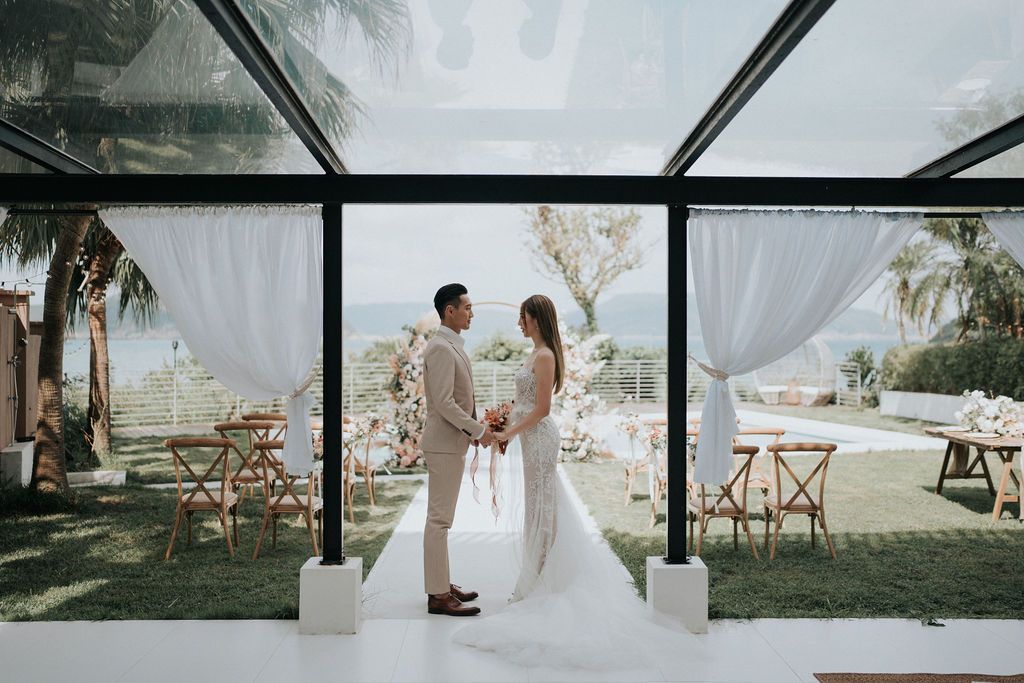 Couple holding hands, getting married under a glass roof. Outdoor ceremony with ocean view, white drapery, and seating.