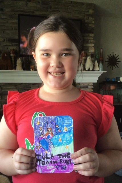 A young girl in a red shirt is holding a card that says tula the tooth fairy