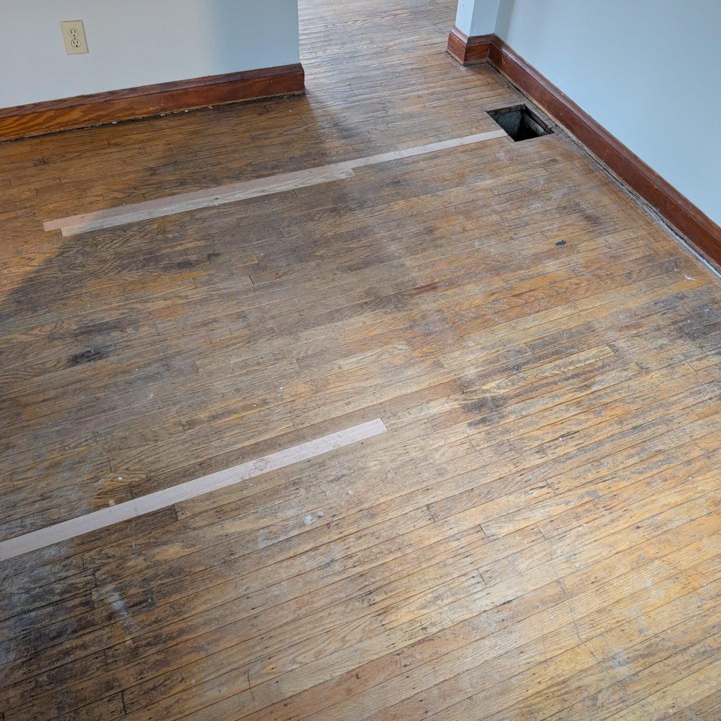 Empty room with worn hardwood floor, white walls, and exposed floor vents.