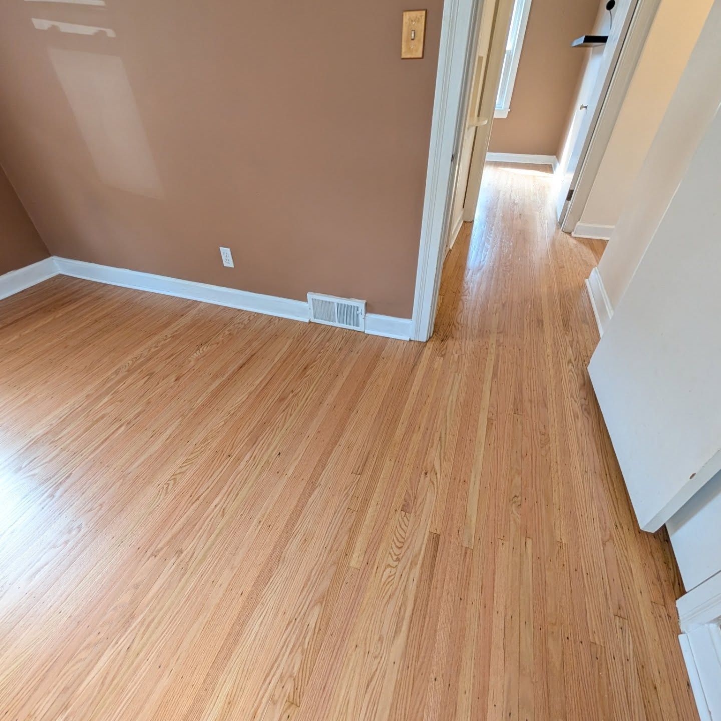Empty hallway with light wood flooring and tan walls, viewed from an adjoining room.