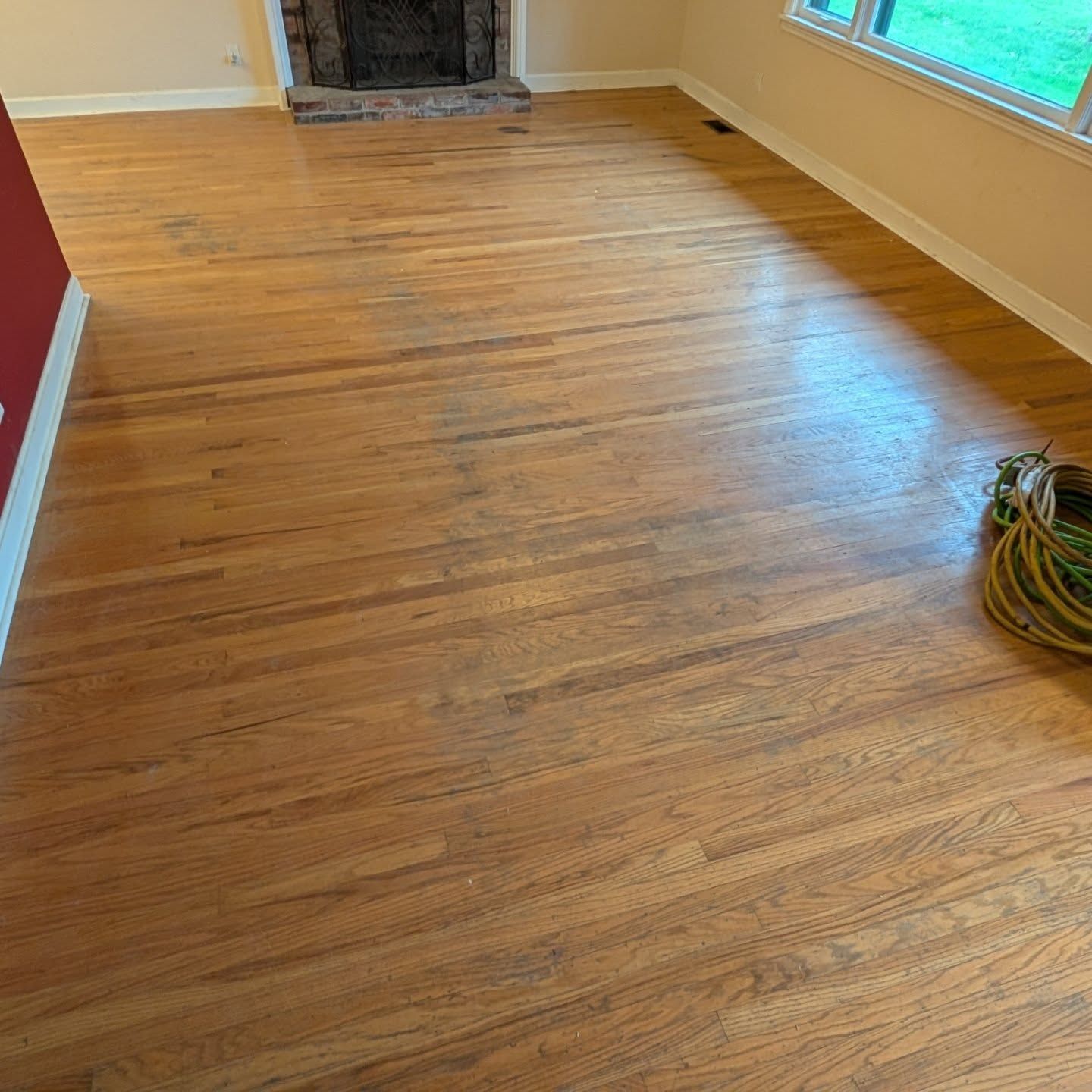 Empty room with newly refinished hardwood floor, fireplace, windows, and a coiled hose on the right.