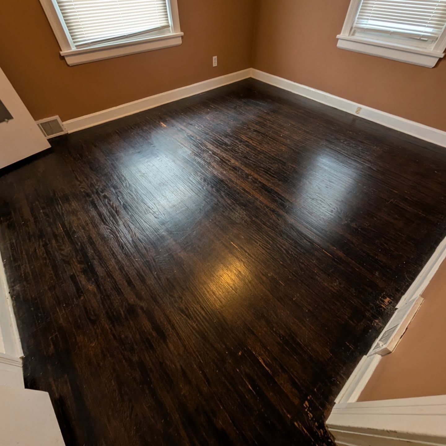 Empty room with dark hardwood floors, tan walls, and two windows with blinds.