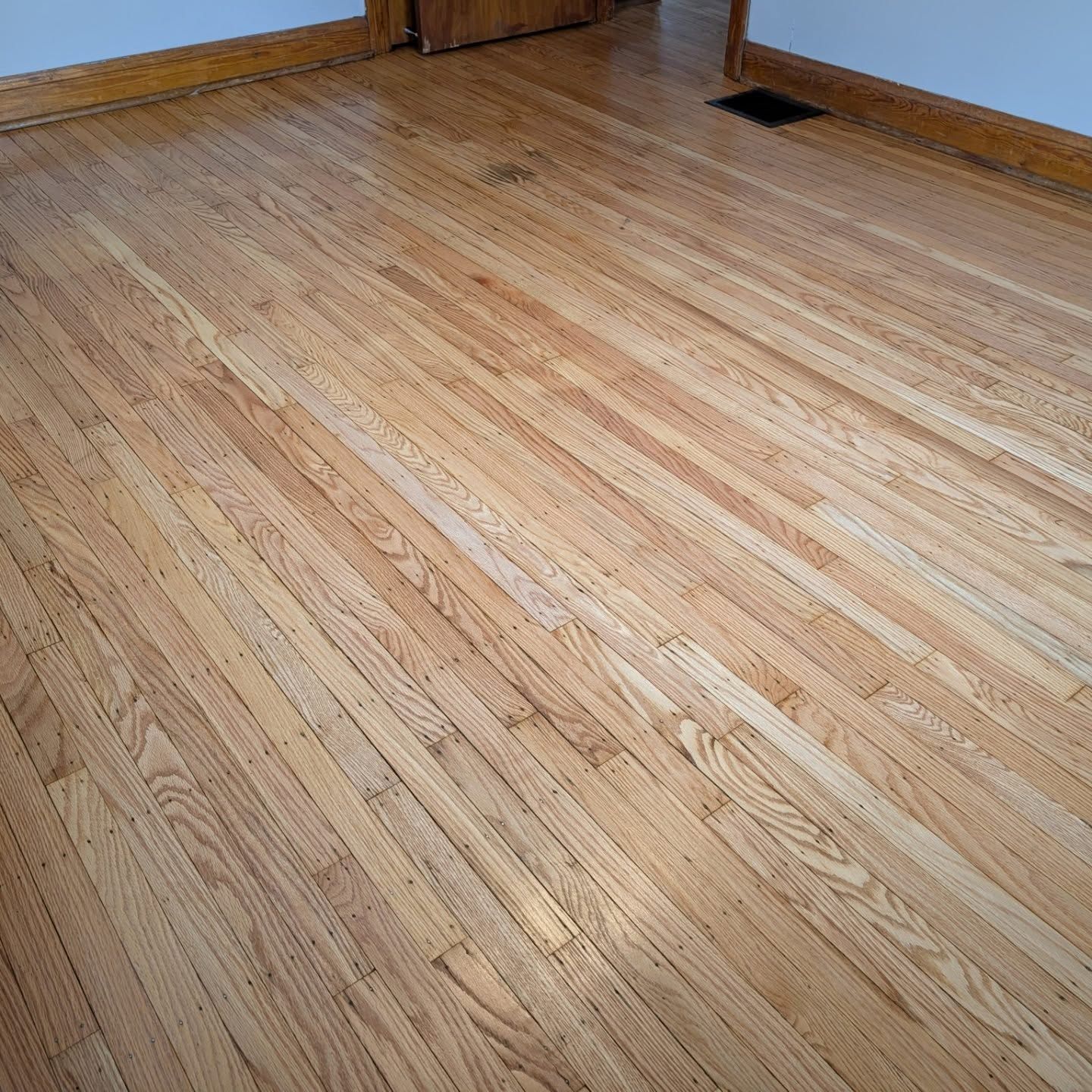 Empty hardwood floor in a room corner with white baseboards and a floor vent.