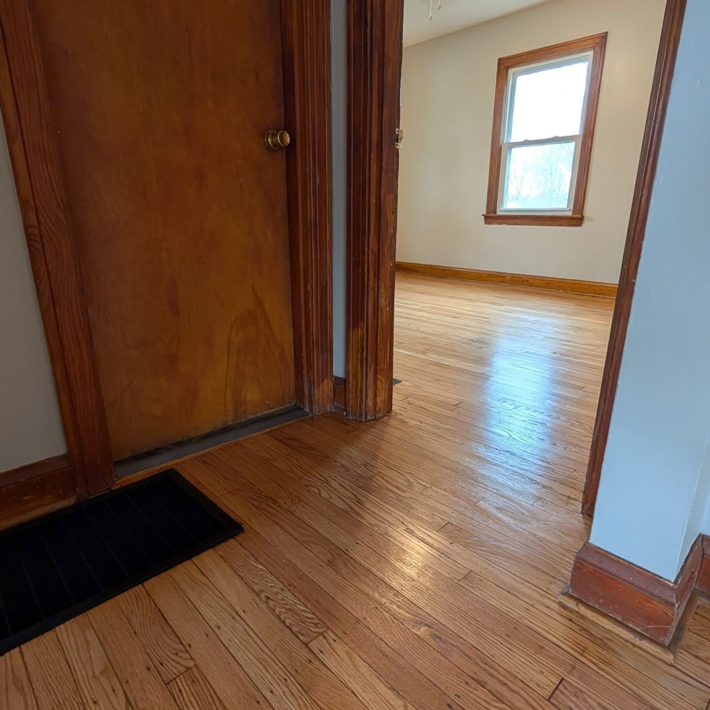 Empty hardwood hallway with wood trim, a black rug, and a bright room visible through an open doorway.