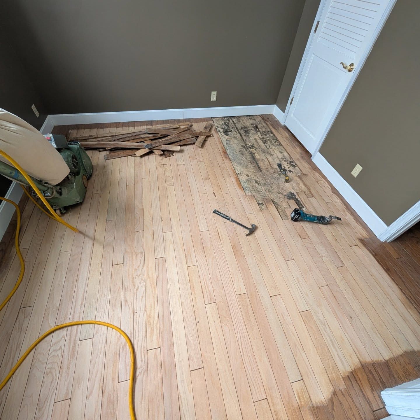 Partially removed hardwood floor in a room with tools and exposed subfloor near the wall