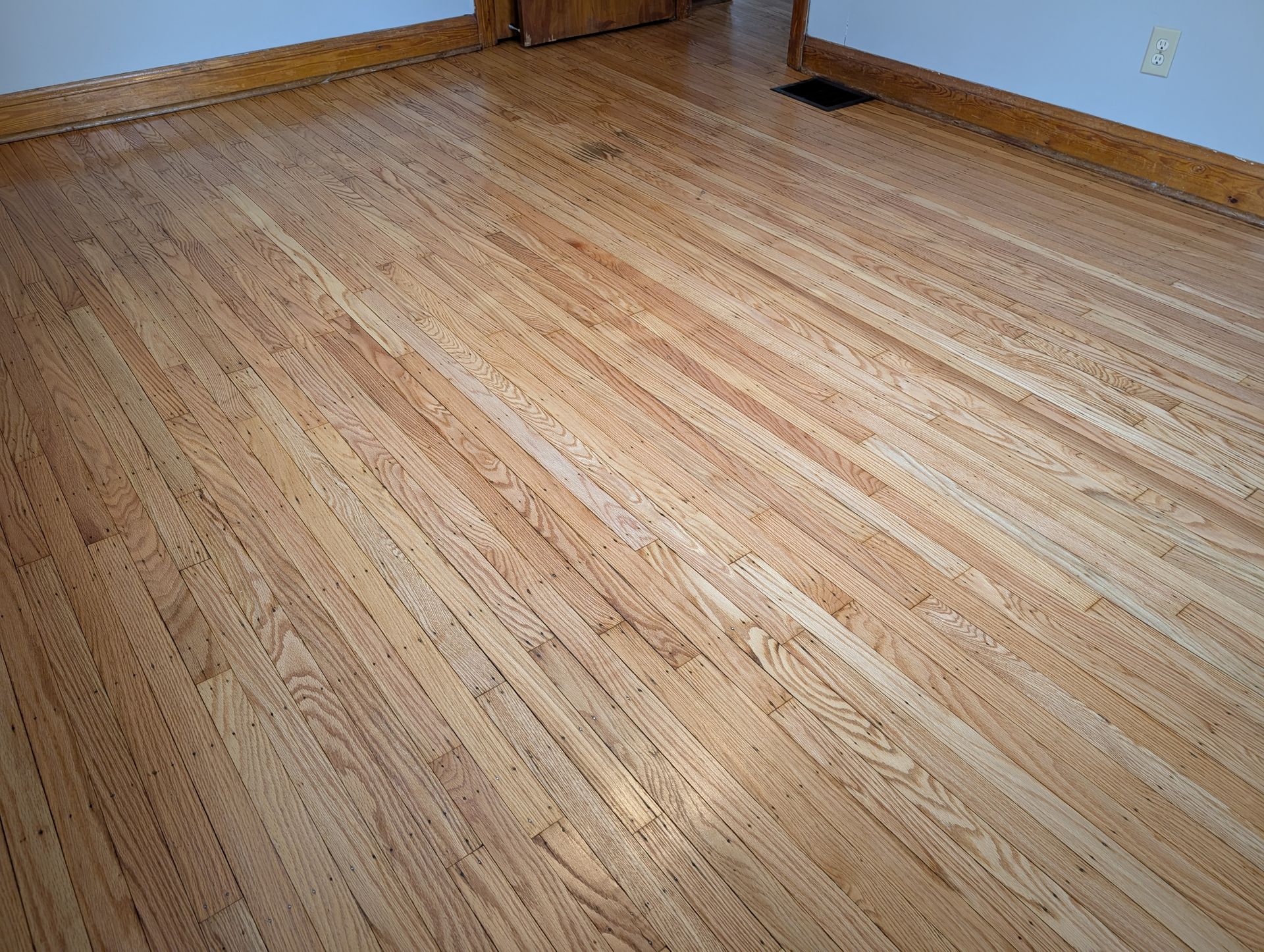 Empty hardwood floor with light wood walls and a floor vent in the corner.