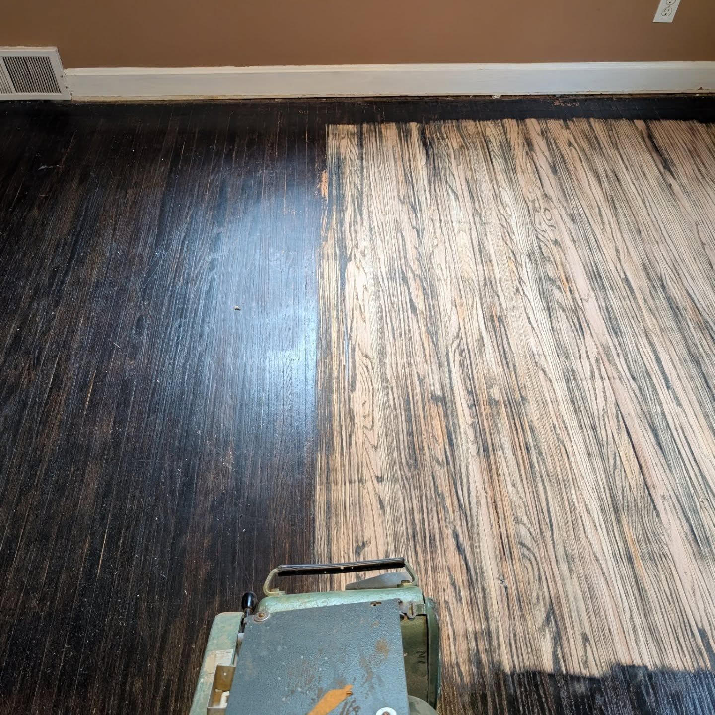 Partially refinished dark wood floor beside lighter striped flooring, with a sanding machine at the bottom edge
