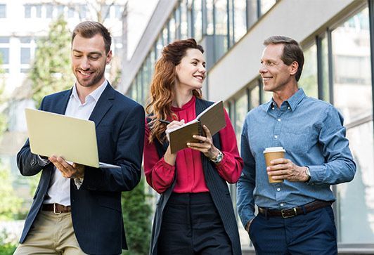 Three office workers walking outside; man with laptop smiles, woman takes notes, man holds coffee.