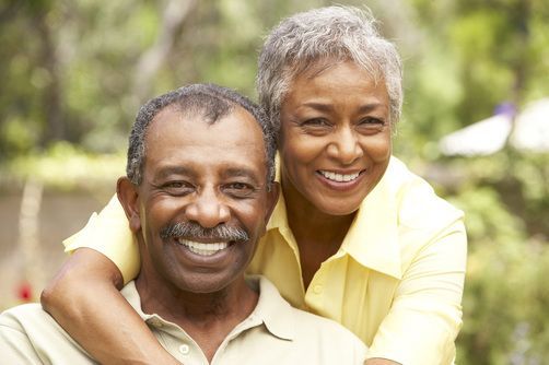 Smiling elderly couple embracing outdoors, woman's arms around man's shoulders, both in yellow shirts.