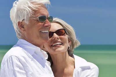 Smiling older couple in sunglasses, embracing on a beach with blue sky.