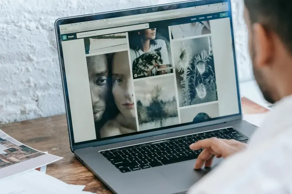 Une personne utilise un ordinateur portable affichant une galerie de photos sur un bureau près d'un mur de briques blanches.