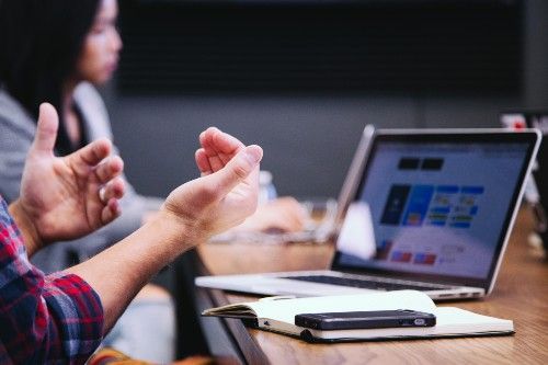 Personne faisant des gestes pendant une réunion, à côté d'un ordinateur portable et d'un carnet posés sur une table de conférence.