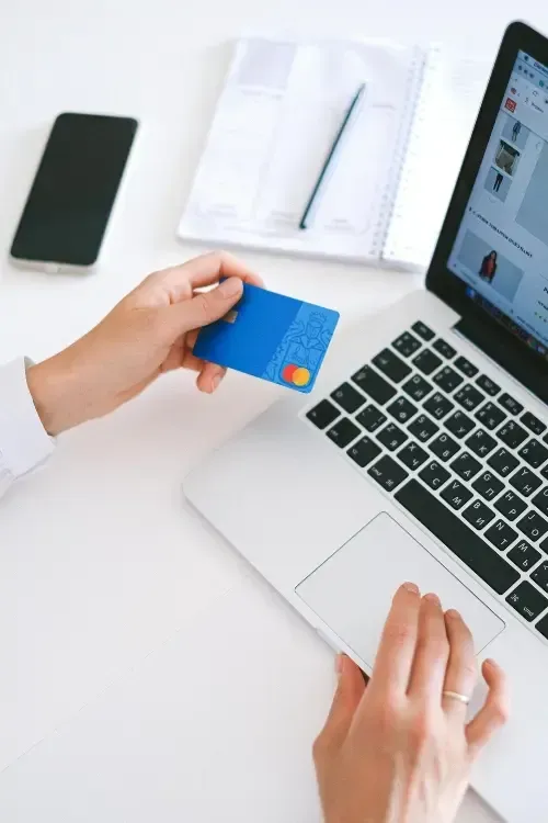 Main tenant une carte de crédit bleue près de l'ordinateur portable pour faire des achats en ligne, avec un carnet et un téléphone sur le bureau
