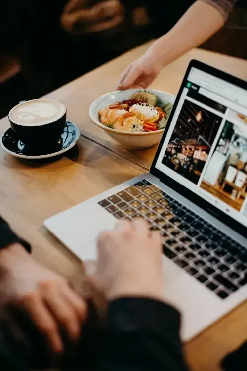 Une personne utilise un ordinateur portable à la table d'un café, avec un café et un bol de nourriture à proximité.