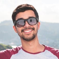 Une personne souriante portant des lunettes de soleil et une chemise bordeaux et blanche se trouve à l'extérieur, avec des montagnes en arrière-plan.