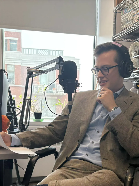 Une personne portant un casque audio et un costume en tweed est assise devant un micro de podcast dans un studio lumineux.