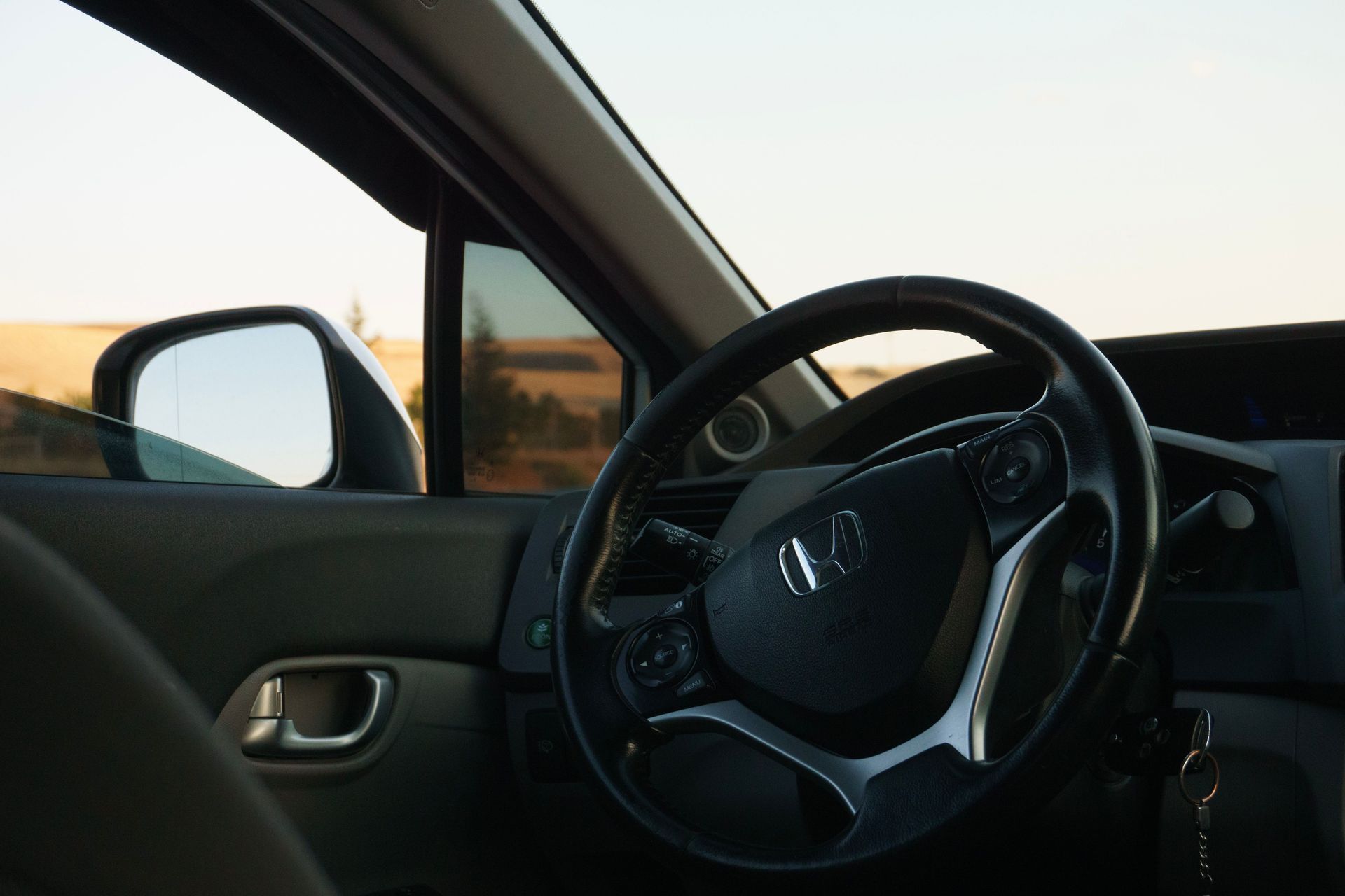 Car interior with steering wheel and side mirror, viewed from the driver’s seat at sunset