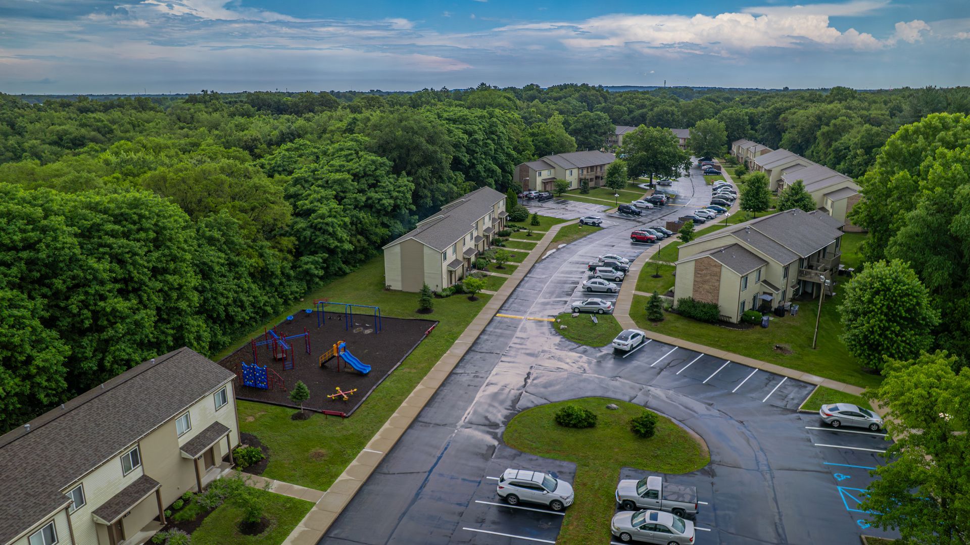 Aerial view of apartment buildings, playground, and parking lot surrounded by green trees.