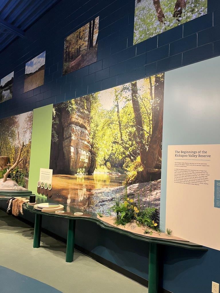 Museum display with landscape photos, a long table with exhibits, and teal walls.