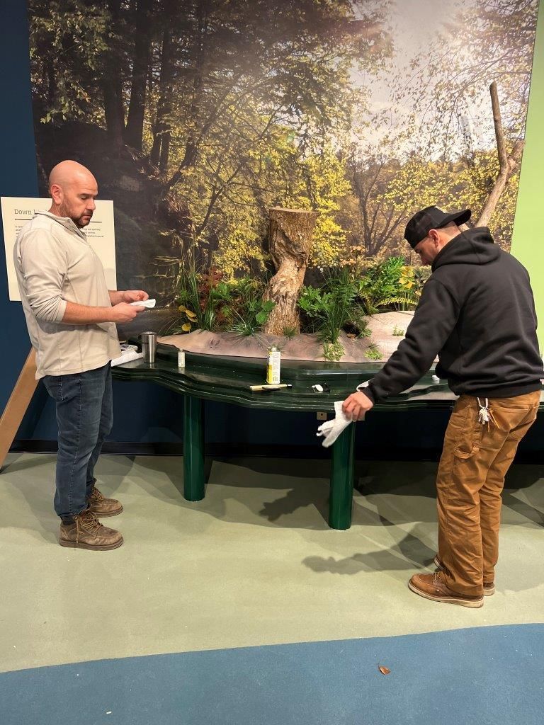Two people working on a diorama with a forest backdrop. One holds papers, the other reaches for materials on the table.