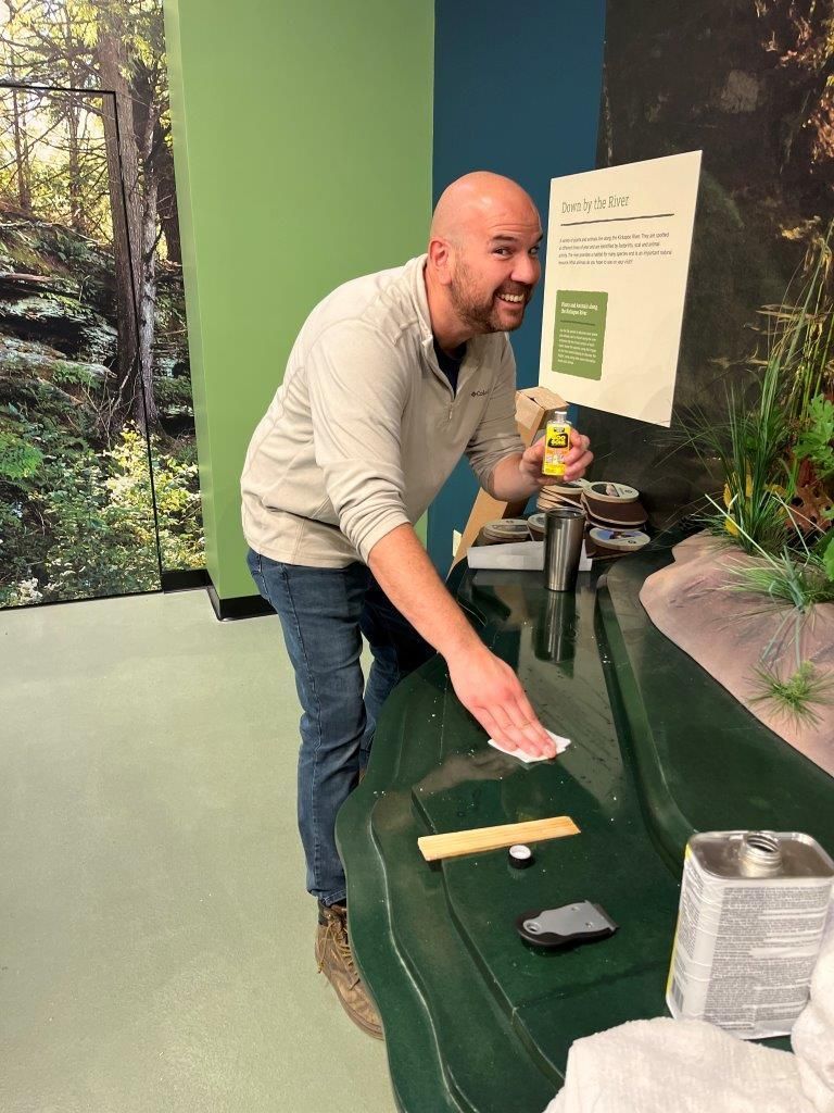 Man cleans a green display surface in a museum, smiling. A bottle and other objects are on the display.