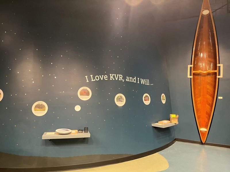 Interior museum display with a cross-section of a canoe on a blue wall. Text 