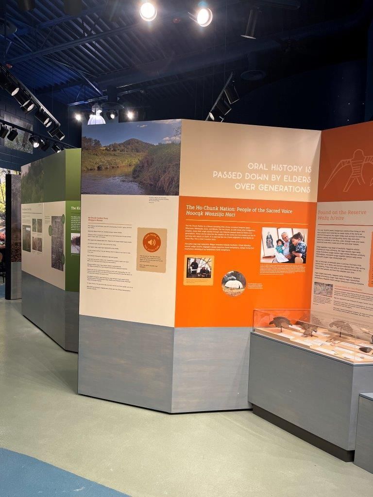 Museum exhibit with information panels featuring photos and text. Panels have green, orange, and tan sections.