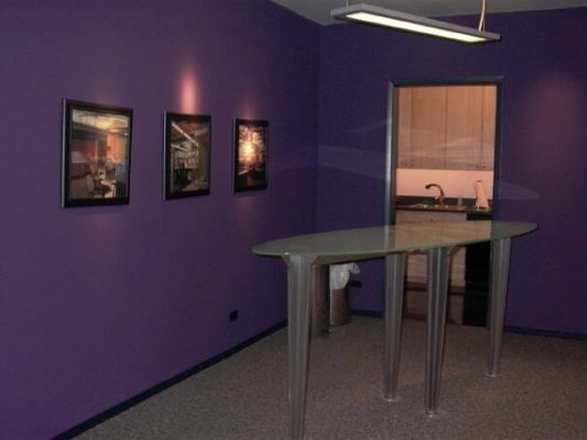 Purple-walled room with a bar, three framed art pieces, and an open doorway to a kitchen area.