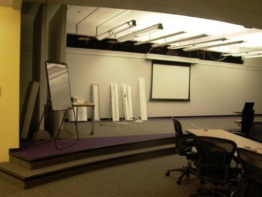 A classroom with a stage, whiteboard, projection screen, desks, and chairs. 