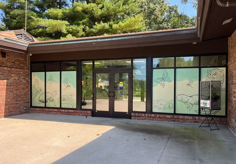 Brick building with large windows, double doors, and decorative decals. Brown trim and a paved patio are in view.