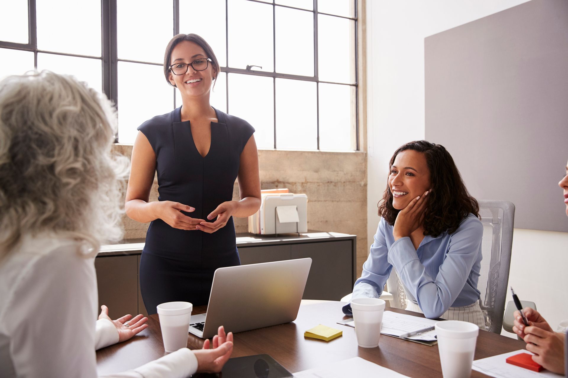 Woman presenting to colleagues in a modern office, smiling; others listen at a table with a laptop.