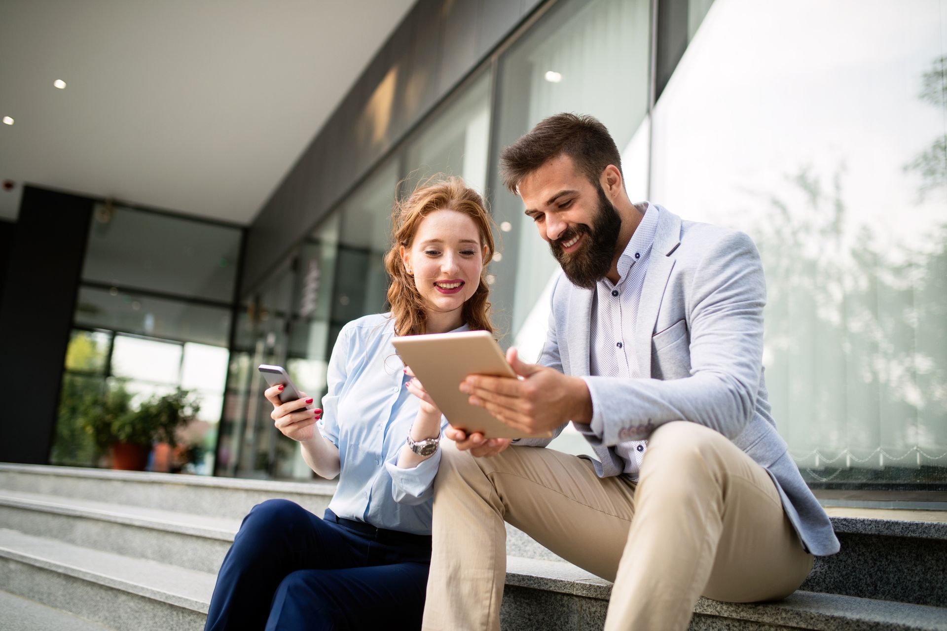 Woman with red hair and man with beard looking at tablet outside a building.
