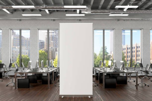 Blank white banner centered in a modern office with desks, computers, and large windows.