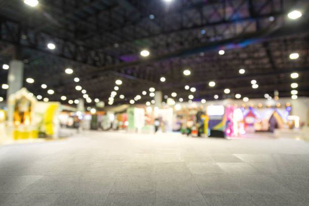 Blurred view of a large convention hall with booths and bright overhead lights.